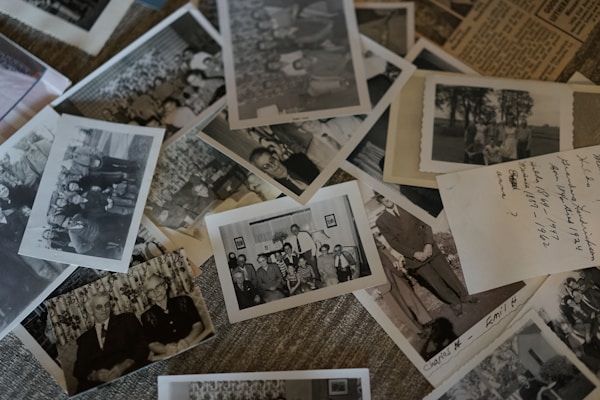 A collection of vintage black and white photographs scattered over a surface, some containing groups of people posing outdoors and others featuring formal portraits. Handwritten notes are visible on some photographs, suggesting dates and names.