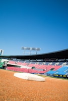 A baseball field with a base prominently in the foreground. The stadium seating in red and blue is visible in the background under a clear blue sky. Tall floodlights tower over the stands.