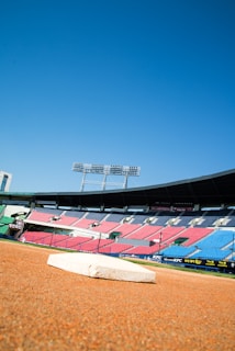 A baseball field with a base prominently in the foreground. The stadium seating in red and blue is visible in the background under a clear blue sky. Tall floodlights tower over the stands.
