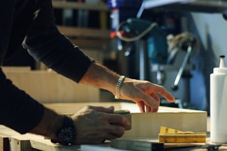 A person is working with wood, wearing a dark long-sleeve shirt and a watch. Hands are focused on sanding or crafting a wooden piece. Various tools and a bottle are visible in the background, suggesting a workshop setting.