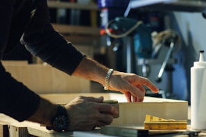 A person is working with wood, wearing a dark long-sleeve shirt and a watch. Hands are focused on sanding or crafting a wooden piece. Various tools and a bottle are visible in the background, suggesting a workshop setting.