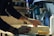 A person is working with wood, wearing a dark long-sleeve shirt and a watch. Hands are focused on sanding or crafting a wooden piece. Various tools and a bottle are visible in the background, suggesting a workshop setting.