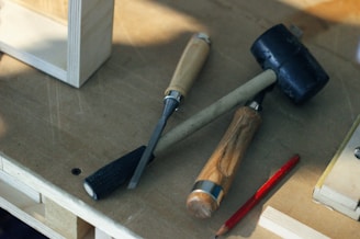 On a wooden surface, there is a rubber mallet, a chisel, a gouge, and a red pencil. The tools are arranged casually, suggesting a workspace environment. The mallet has a black rubber head with a wooden handle, while both the chisel and gouge have wooden handles and metal blades. The red pencil is unsharpened and placed beside the tools.
