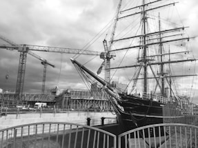 A large ship with multiple masts, set in a dockyard environment surrounded by construction cranes. Scaffolding and a structured building site are visible in the background. The image has a monochromatic filter, creating a classic, historical feel.