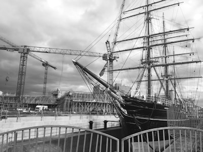 A large ship with multiple masts, set in a dockyard environment surrounded by construction cranes. Scaffolding and a structured building site are visible in the background. The image has a monochromatic filter, creating a classic, historical feel.