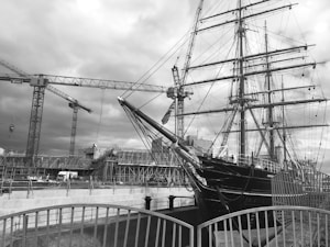 A large ship with multiple masts, set in a dockyard environment surrounded by construction cranes. Scaffolding and a structured building site are visible in the background. The image has a monochromatic filter, creating a classic, historical feel.
