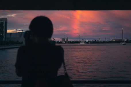 A silhouette of a person holding a camera and looking out over a body of water, with a dramatic sunset in the background. The sky is filled with vibrant shades of pink and purple, and industrial structures can be seen across the water.