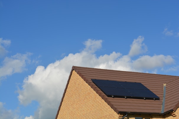 A sloped roof with dark solar panels against a bright blue sky with scattered white clouds. The roof is covered with brown tiles and the walls are made of light-colored bricks.