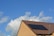 A sloped roof with dark solar panels against a bright blue sky with scattered white clouds. The roof is covered with brown tiles and the walls are made of light-colored bricks.