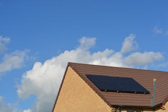 A sloped roof with dark solar panels against a bright blue sky with scattered white clouds. The roof is covered with brown tiles and the walls are made of light-colored bricks.