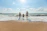 A family of three stands on a sandy beach, facing the ocean waves. The sky is clear with a few scattered clouds, and the sunlight casts a bright glow across the scene.