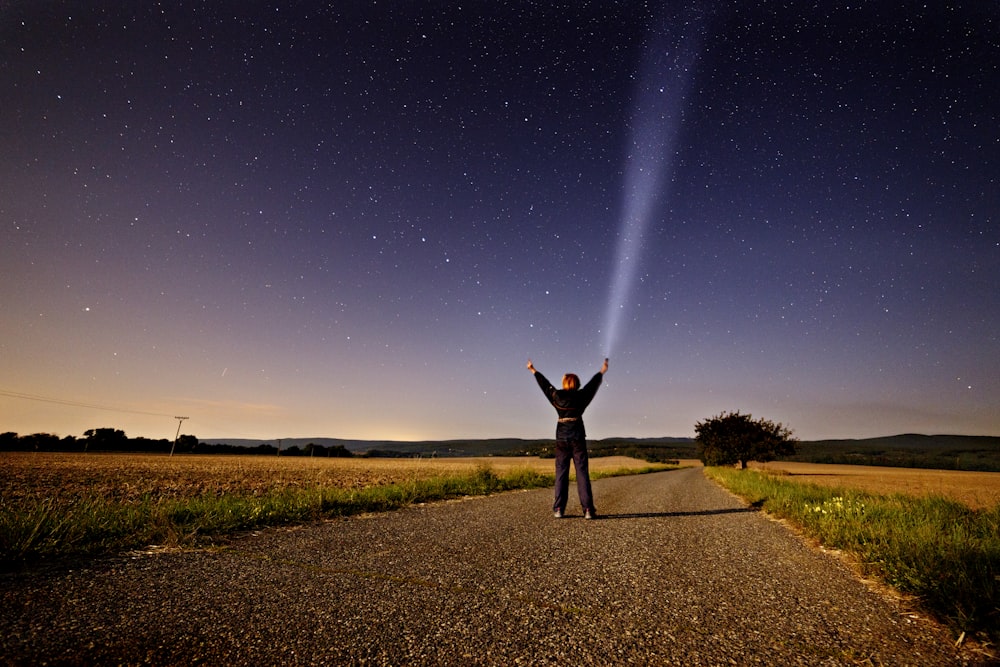 person raising his two hands while holding flashlight standing on road between grass during sunset