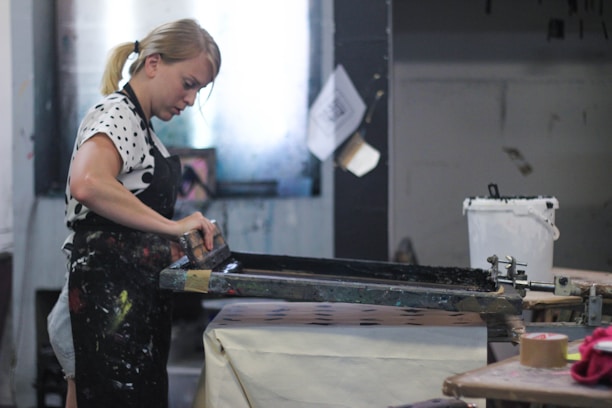 A person is engaged in a screen printing process, using a squeegee to apply ink across a screen onto a surface below. The individual is wearing a polka-dotted shirt and dark apron with paint stains, indicating an artistic environment. The setting includes a workbench, a container, and other tools related to the printing process.
