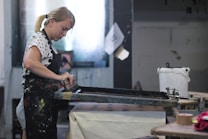 A person is engaged in a screen printing process, using a squeegee to apply ink across a screen onto a surface below. The individual is wearing a polka-dotted shirt and dark apron with paint stains, indicating an artistic environment. The setting includes a workbench, a container, and other tools related to the printing process.