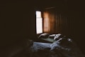 Pale light pouring into a cabin through a small window illuminates two unmade beds