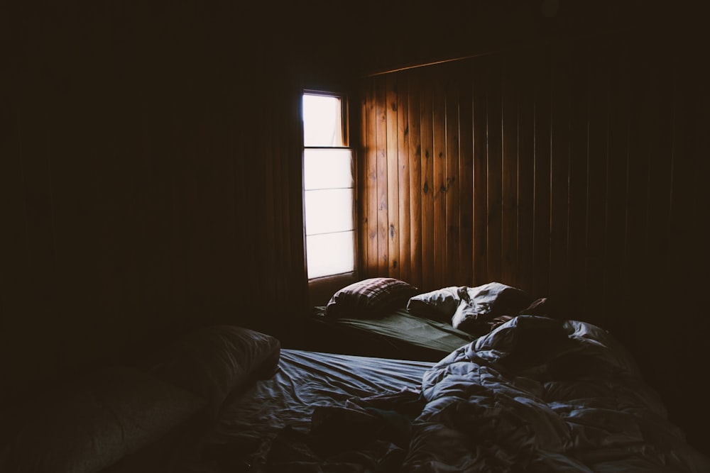 Pale light pouring into a cabin through a small window illuminates two unmade beds