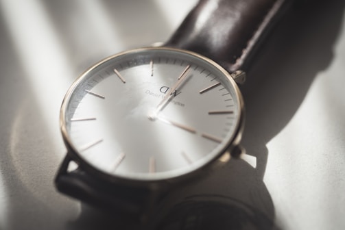 A close-up view of a minimalist wristwatch with a white face, slender hour markers, and a simple design. The watch has a black leather strap and is captured in soft lighting that casts gentle shadows.