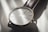 A close-up view of a minimalist wristwatch with a white face, slender hour markers, and a simple design. The watch has a black leather strap and is captured in soft lighting that casts gentle shadows.