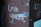 Graffiti on a wall with the message 'Love yourself' written in white paint, accompanied by a small heart symbol. The wall has a rough texture, with visible screws and some peeling patches of paint and tape.
