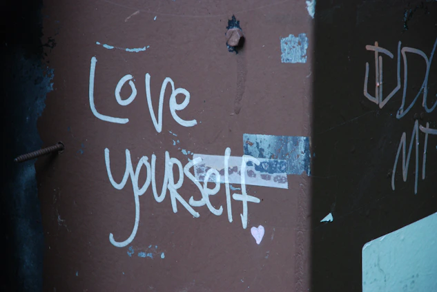 Graffiti on a wall with the message 'Love yourself' written in white paint, accompanied by a small heart symbol. The wall has a rough texture, with visible screws and some peeling patches of paint and tape.