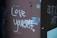 Graffiti on a wall with the message 'Love yourself' written in white paint, accompanied by a small heart symbol. The wall has a rough texture, with visible screws and some peeling patches of paint and tape.