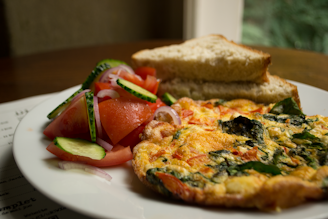 A plate of food featuring a colorful omelette filled with green vegetables and red tomatoes, accompanied by a side salad of fresh tomatoes, cucumbers, and onion slices. Two pieces of bread are stacked behind the omelette on the plate. The setting is a cozy indoor environment with natural light coming through a nearby window.