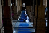 Several individuals are seated on blue prayer rugs amid a row of columns inside a dimly lit religious building. They appear to be engaged in reading or prayer, dressed in white robes and caps, creating a serene and contemplative atmosphere. The architecture features stone columns and decorative windows in the background, suggesting a historical or culturally significant setting.
