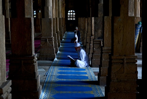 Several individuals are seated on blue prayer rugs amid a row of columns inside a dimly lit religious building. They appear to be engaged in reading or prayer, dressed in white robes and caps, creating a serene and contemplative atmosphere. The architecture features stone columns and decorative windows in the background, suggesting a historical or culturally significant setting.