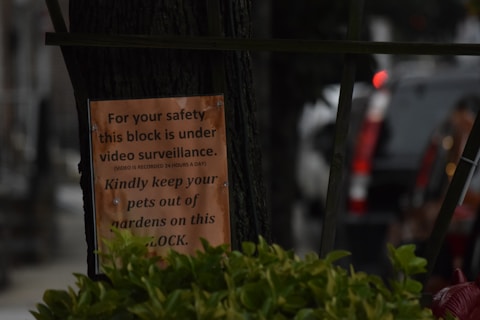 A sign is attached to a tree, warning residents about video surveillance and advising them to keep pets out of gardens. The background features parked cars and blurred urban elements, with some greenery visible in the foreground.