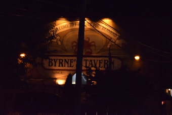 A dimly lit sign of a tavern is visible, with ornate text and a decorative element above it. The background is dark, with a few small bright lights accentuating the sign. Silhouettes of trees and electrical wires can be seen.