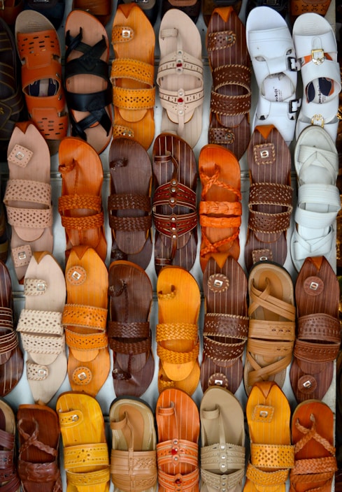 A variety of leather sandals in different designs and shades of brown, orange, and white are neatly arranged in rows. The sandals feature different patterns and textures, showcasing craftsmanship and diversity in style.