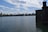 The image features a large body of calm water with the New York City skyline visible in the background. The sky is partly cloudy, and there's a stone structure on the right side of the image. A fountain can be seen in the distance, and a few birds are flying above.