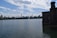 The image features a large body of calm water with the New York City skyline visible in the background. The sky is partly cloudy, and there's a stone structure on the right side of the image. A fountain can be seen in the distance, and a few birds are flying above.