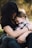 A nurturing embrace between an adult and a child, conveying warmth and comfort. The adult holds the child close, as the child nestles their head in the adult's chest. The image is softly focused with a natural outdoor background blurred behind them.