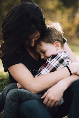 A nurturing embrace between an adult and a child, conveying warmth and comfort. The adult holds the child close, as the child nestles their head in the adult\'s chest. The image is softly focused with a natural outdoor background blurred behind them.