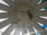 A large, circular ceiling adorned with geometric window panels that allow natural light to filter through. Suspended sculptures of angels can be seen, creating a sense of movement and grace. The architectural design features stylized rays extending outward, enhancing the spiritual and serene atmosphere.
