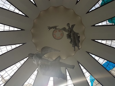 A large, circular ceiling adorned with geometric window panels that allow natural light to filter through. Suspended sculptures of angels can be seen, creating a sense of movement and grace. The architectural design features stylized rays extending outward, enhancing the spiritual and serene atmosphere.