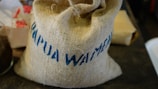 A burlap sack with blue text prominently displayed, indicating 'Papua Waimena.' Nearby, there are various blurry items, including a white object with a red floral pattern and possibly a glass container.
