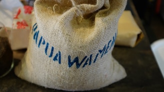 A burlap sack with blue text prominently displayed, indicating 'Papua Waimena.' Nearby, there are various blurry items, including a white object with a red floral pattern and possibly a glass container.