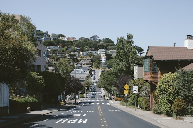 A quiet suburban street with a steep incline, lined with trees and houses. Pedestrian signs are visible on the right, while the distant landscape is dotted with greenery and more residences. The road markings indicate a speed limit and other traffic instructions.