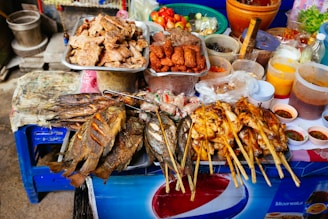 An assortment of grilled and fried foods displayed on a street food stall. There are fried fish, skewers of grilled chicken, and various other meats on plates. Surrounding the main dishes, there are containers with sauces, fresh vegetables like tomatoes and lettuce, and trays of condiments. The vibrant colors suggest a traditional and busy market atmosphere.