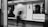 A black and white photograph of a subway station platform with a blurred figure walking by. Two large movie posters are prominently displayed on a tiled wall, featuring close-up portraits. The posters hint at a cinematic theme with text possibly indicative of movie titles or reviews. The scene is captured from within a train carriage, with parts of the doorway visible.