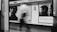 A black and white photograph of a subway station platform with a blurred figure walking by. Two large movie posters are prominently displayed on a tiled wall, featuring close-up portraits. The posters hint at a cinematic theme with text possibly indicative of movie titles or reviews. The scene is captured from within a train carriage, with parts of the doorway visible.