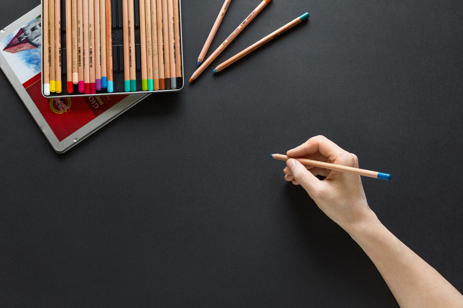 A tin of pencils lined up in a row, with a hand holding one beside it