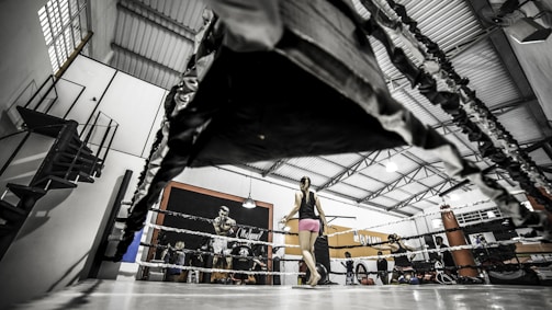 A spacious gym interior featuring a boxing ring is visible. The image is taken from a low angle, emphasizing the ropes of the ring in the foreground. A person stands inside the ring wearing athletic attire, and there are various gym equipment around, including a punching bag and ropes. The walls are decorated with posters, one prominently displaying a boxer.