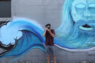 A person is taking a photo with a camera in front of a mural depicting a stylized ocean wave and a face with long hair. The mural is painted on a concrete wall and features vibrant shades of blue and white.