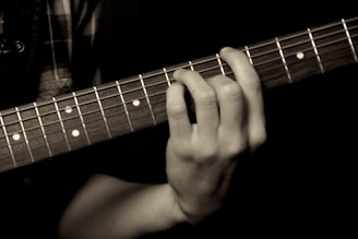 A close-up of a hand playing an acoustic guitar, with fingers pressing down on the strings against the fretboard. The image focuses on the details of the hand and guitar, highlighting the strings and frets.