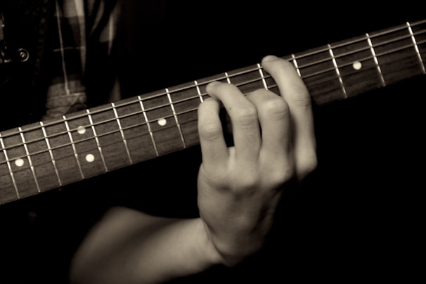 A close-up of a hand playing an acoustic guitar, with fingers pressing down on the strings against the fretboard. The image focuses on the details of the hand and guitar, highlighting the strings and frets.