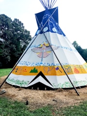 A vividly painted tipi stands in an outdoor setting, surrounded by grass and trees. The tipi features colorful designs, including a large bird motif, abstract shapes, and handprints. The structure is supported by wooden poles, creating a traditional and cultural aesthetic.
