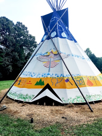 A vividly painted tipi stands in an outdoor setting, surrounded by grass and trees. The tipi features colorful designs, including a large bird motif, abstract shapes, and handprints. The structure is supported by wooden poles, creating a traditional and cultural aesthetic.